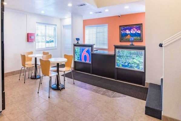 A hotel reception area with a counter, a TV screen displaying the number 6, and a seating area with chairs and tables.