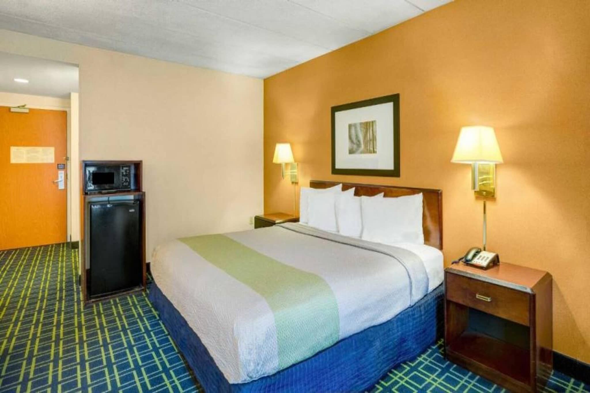 Una habitación de hotel con cama, escritorio, teléfono, lámpara y refrigerador.