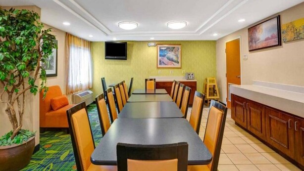 Ein Hotel-Speisesaal mit einem langen Tisch, Stühlen, einem Fernseher, einem Gemälde und einer Topfpflanze.