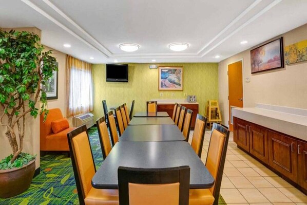 Una sala da pranzo di un hotel con un lungo tavolo, sedie, un televisore, un quadro e una pianta in vaso.