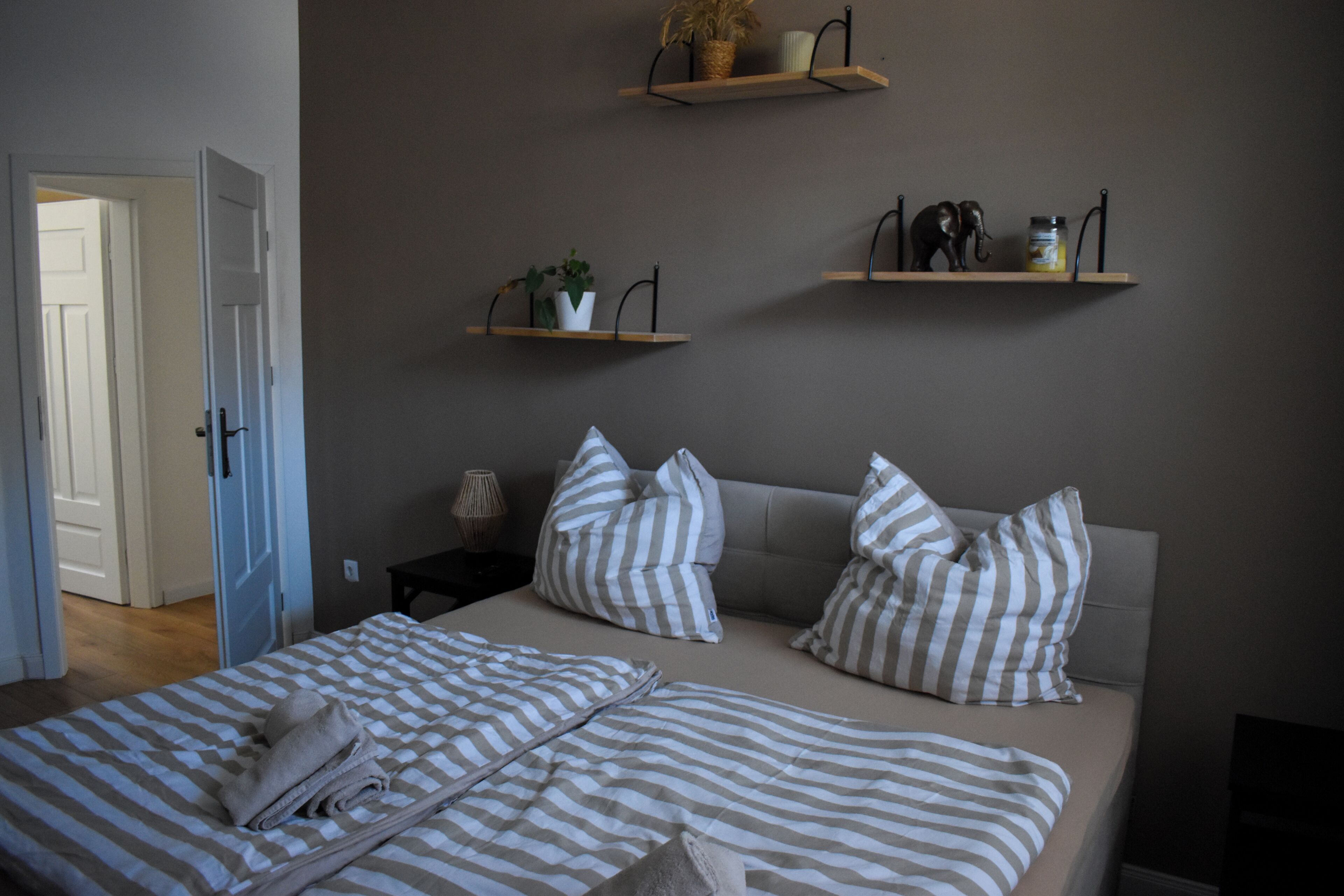 Un dormitorio con cama, dos almohadas, una mesita de noche, una lámpara y dos estantes en la pared con objetos decorativos.