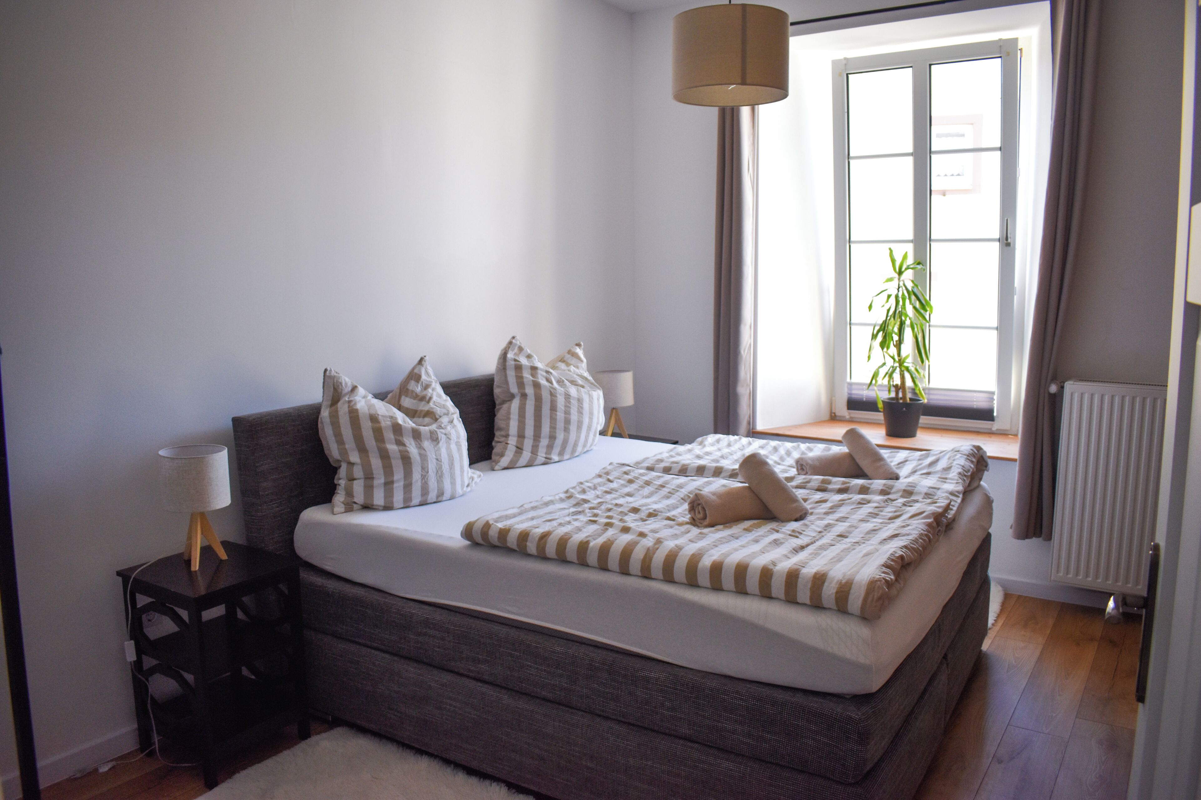 Een slaapkamer met een groot bed, een nachtkastje, een lamp, een raam met gordijnen en een potplant.