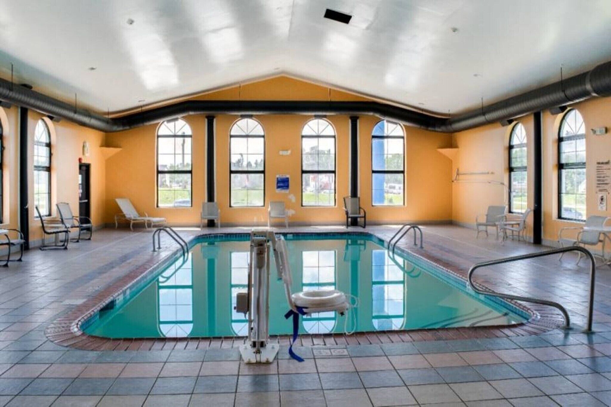An indoor swimming pool with clear glass walls, multiple windows, and lounge chairs.