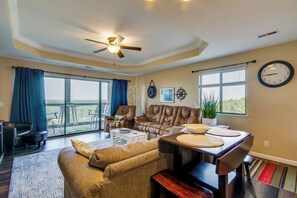 A spacious living room with a large sofa, a coffee table, and a dining area with a table and chairs. There is a ceiling fan, a clock on the wall, and a window with blinds. - 'The Wake Zone' Condo Near Lake of the Ozarks (Lake Ozark)