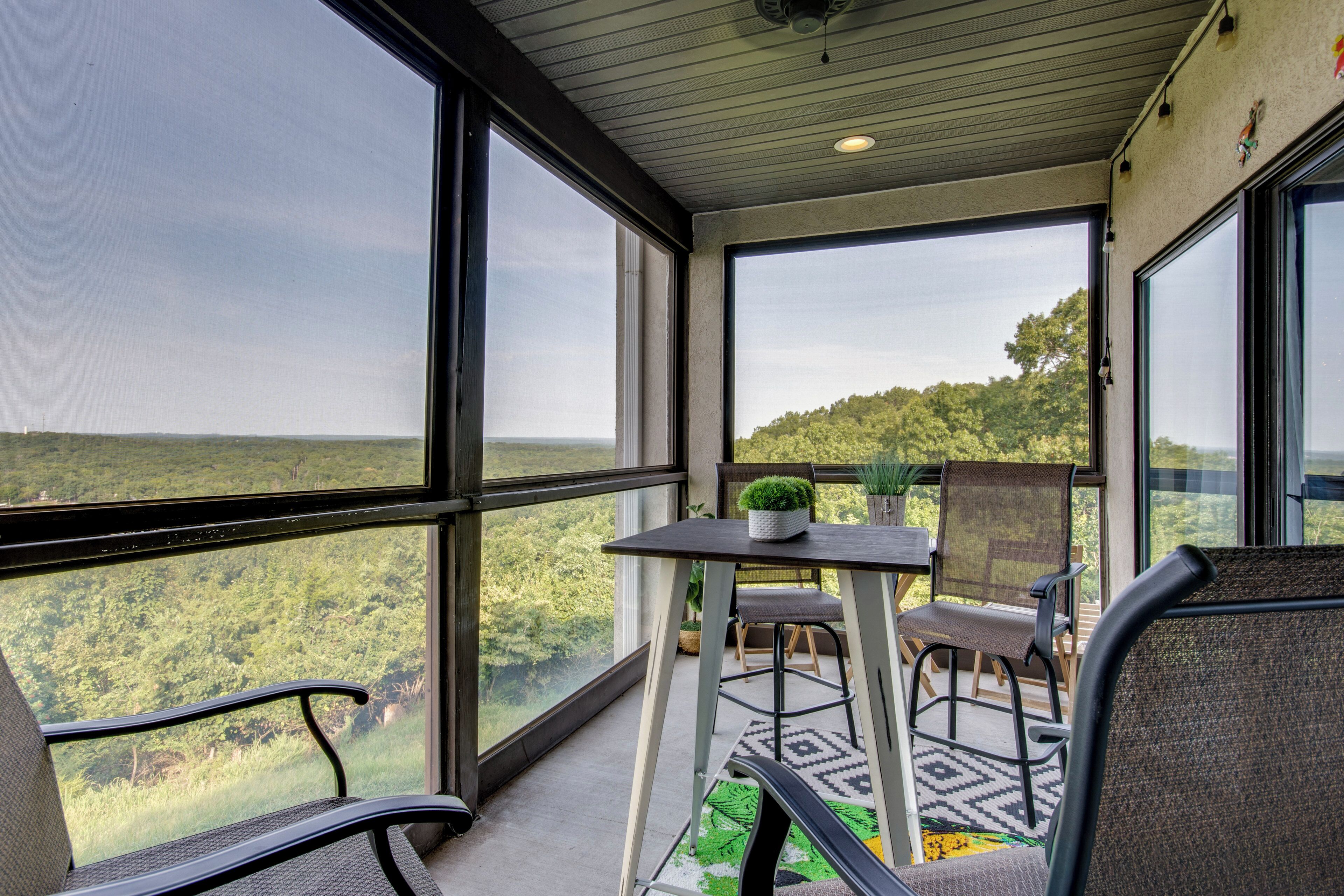 A balcony with a view of a forested landscape, a table with chairs, and a potted plant.