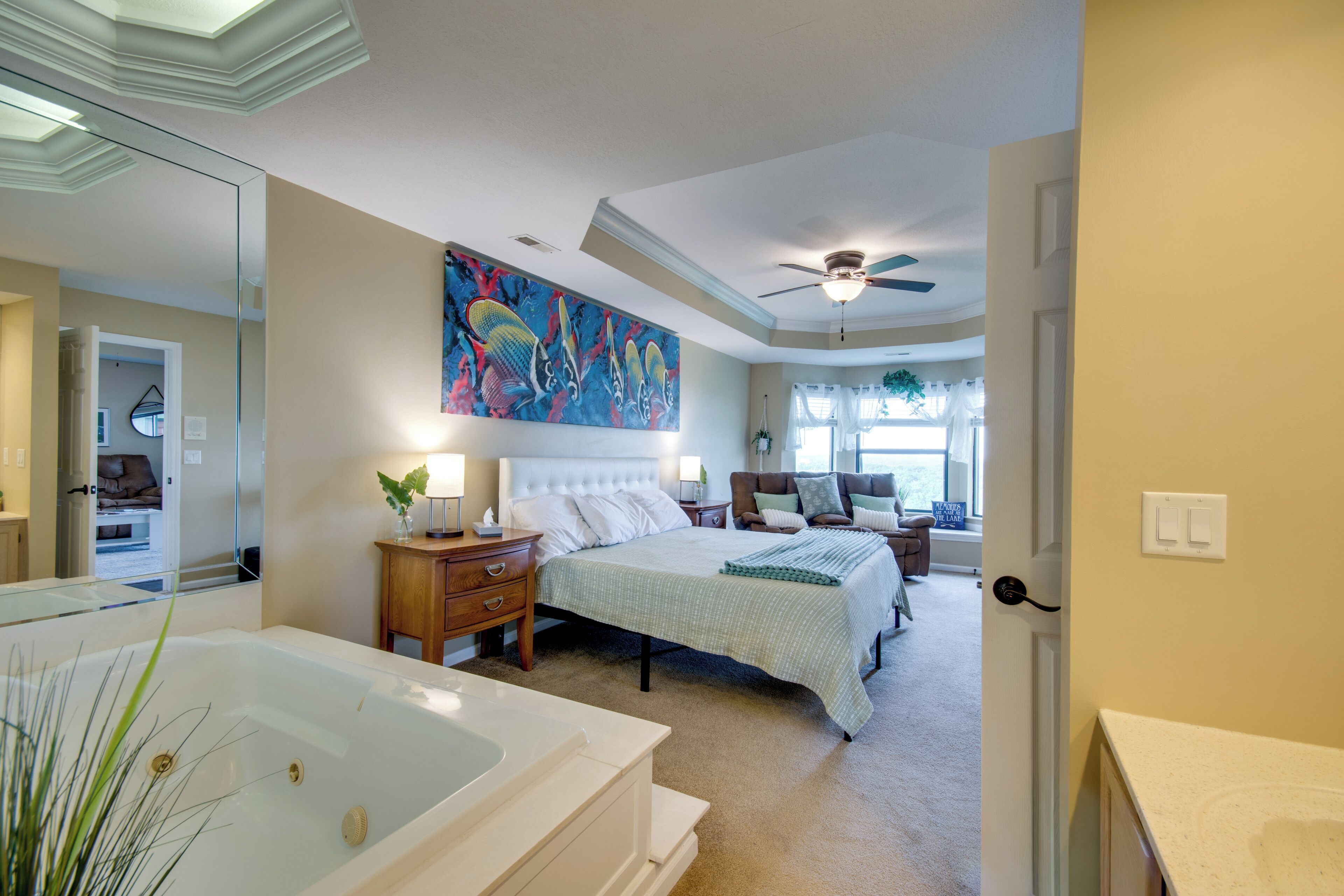 A bedroom with a large bed, a nightstand, a dresser, a ceiling fan, a window with curtains, and a bathroom with a bathtub.
