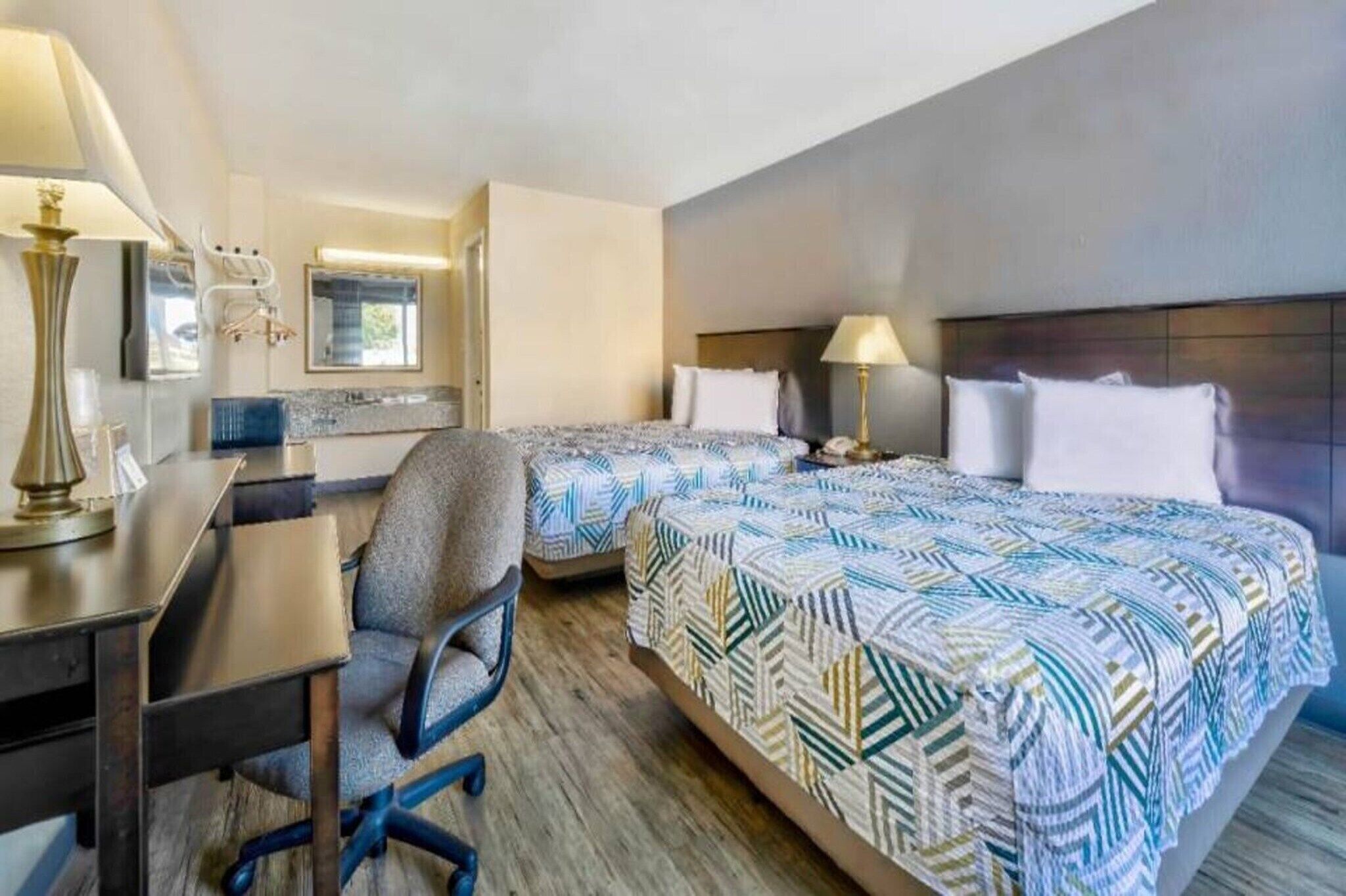 A hotel room with two beds, a desk, a chair, and a lamp.