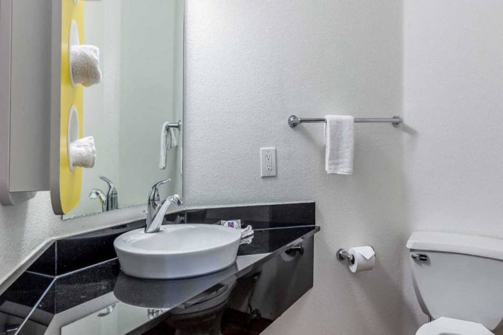 A bathroom with a sink, mirror, towel rack, toilet, and a roll of toilet paper.