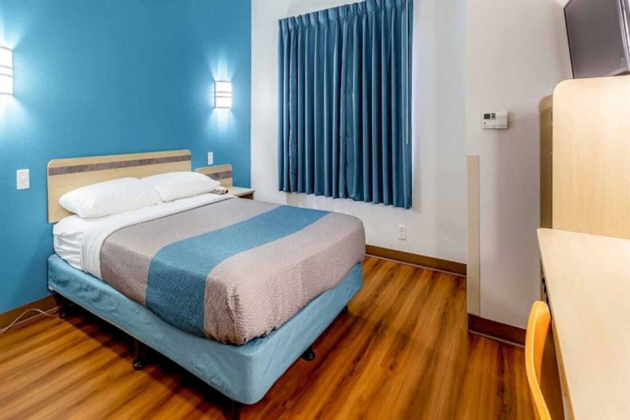 A hotel room with a bed, a window with curtains, a wooden floor, and a chair.
