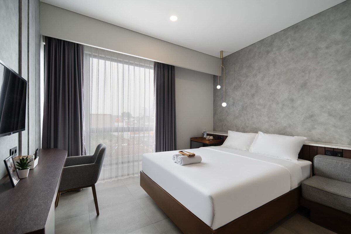 Habitación de hotel moderna con una cama grande, un escritorio, una silla, un televisor y vistas a la ciudad.
