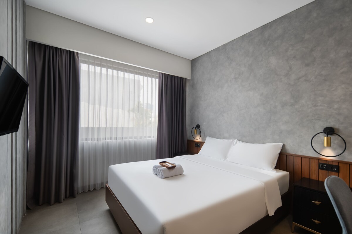 Una habitación de hotel con una cama grande, televisor de pantalla plana, mesita de noche y lámpara de pared.
