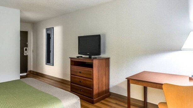 A hotel room with a bed, a wooden dresser, a television, a desk, and a chair.