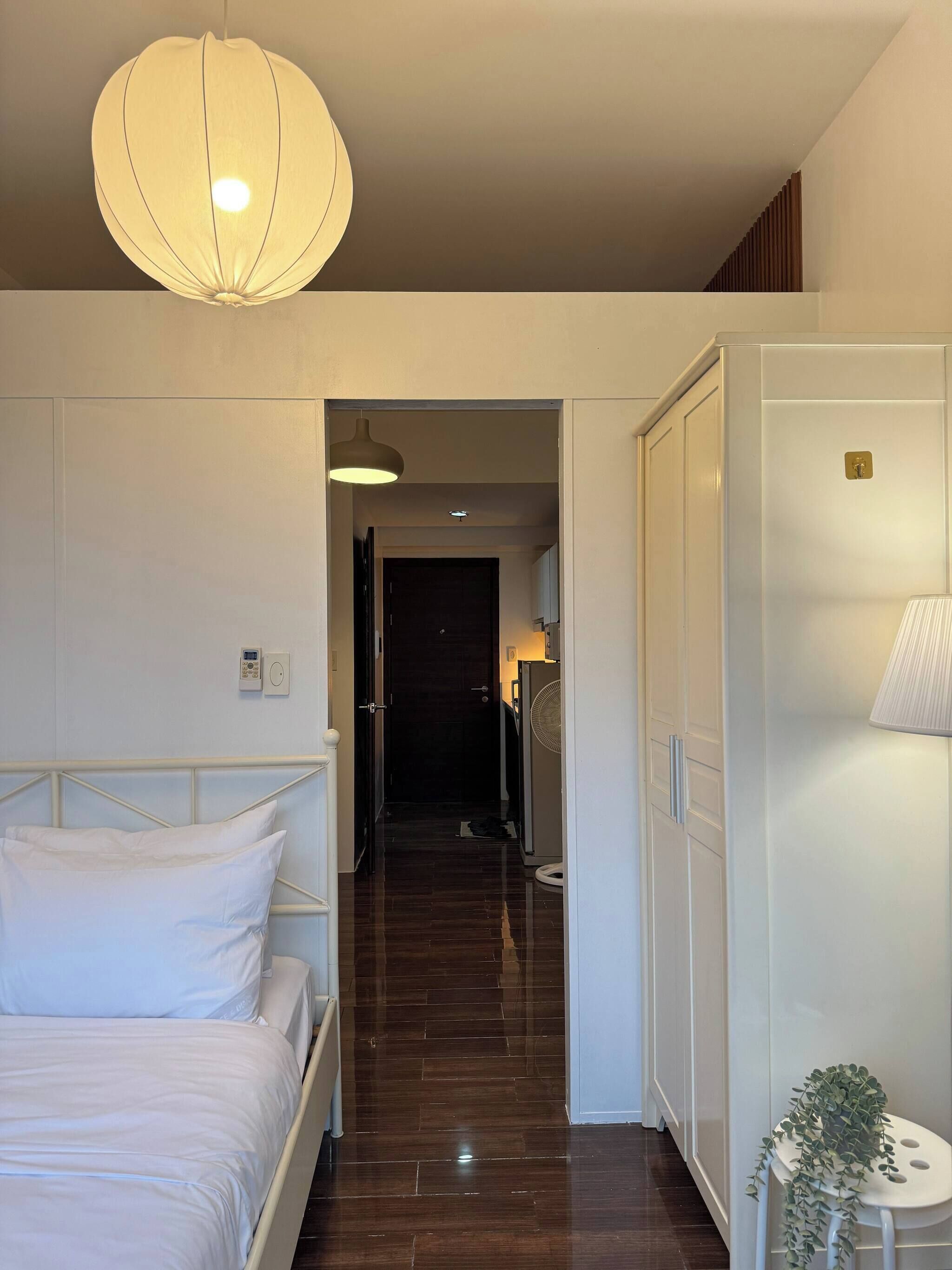 Une chambre d’hôtel avec un lit, une table de chevet avec une lampe, un couloir menant à une porte, et une petite plante posée sur un tabouret.