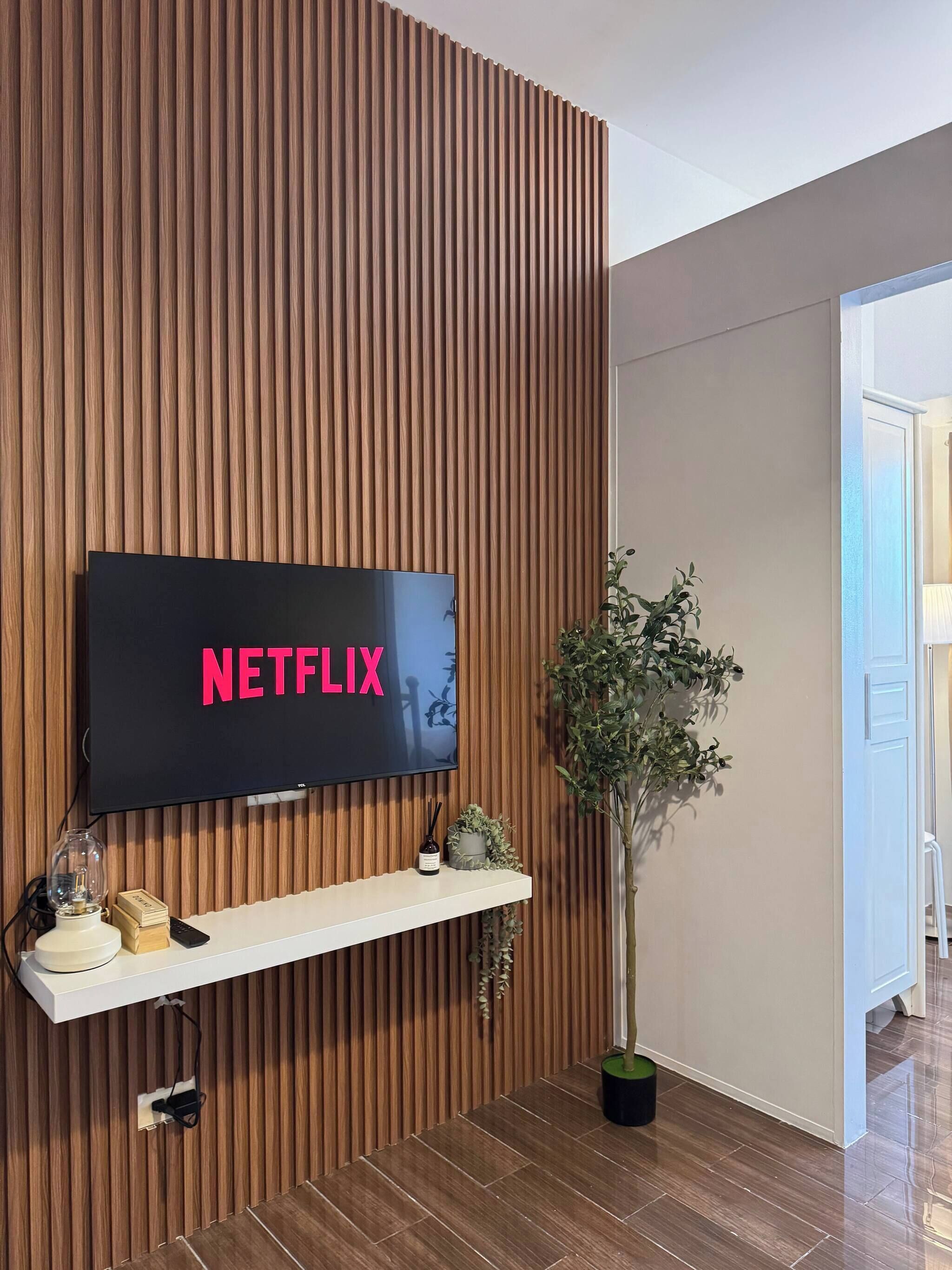 Un salon moderne avec un mur recouvert de panneaux en bois, un téléviseur fixé au mur affichant le logo « NETFLIX », une étagère blanche ornée d’objets décoratifs et une plante en pot.