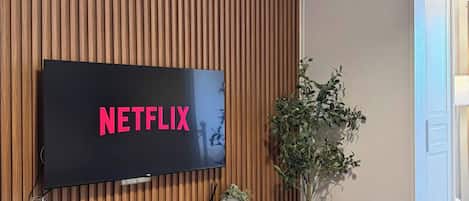 Un salon moderne avec un mur recouvert de panneaux en bois, un téléviseur fixé au mur affichant le logo « NETFLIX », une étagère blanche ornée d’objets décoratifs et une plante en pot.
