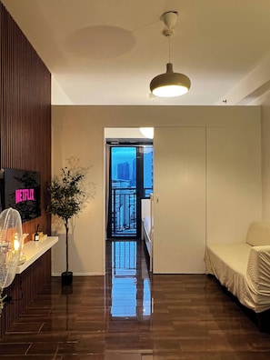 A hotel room with a wooden floor, a bed, a bedside table with a fan, a potted plant, and a TV mounted on the wall. - Central Makati City View with Fast WiFi and Netflix (Makati City)
