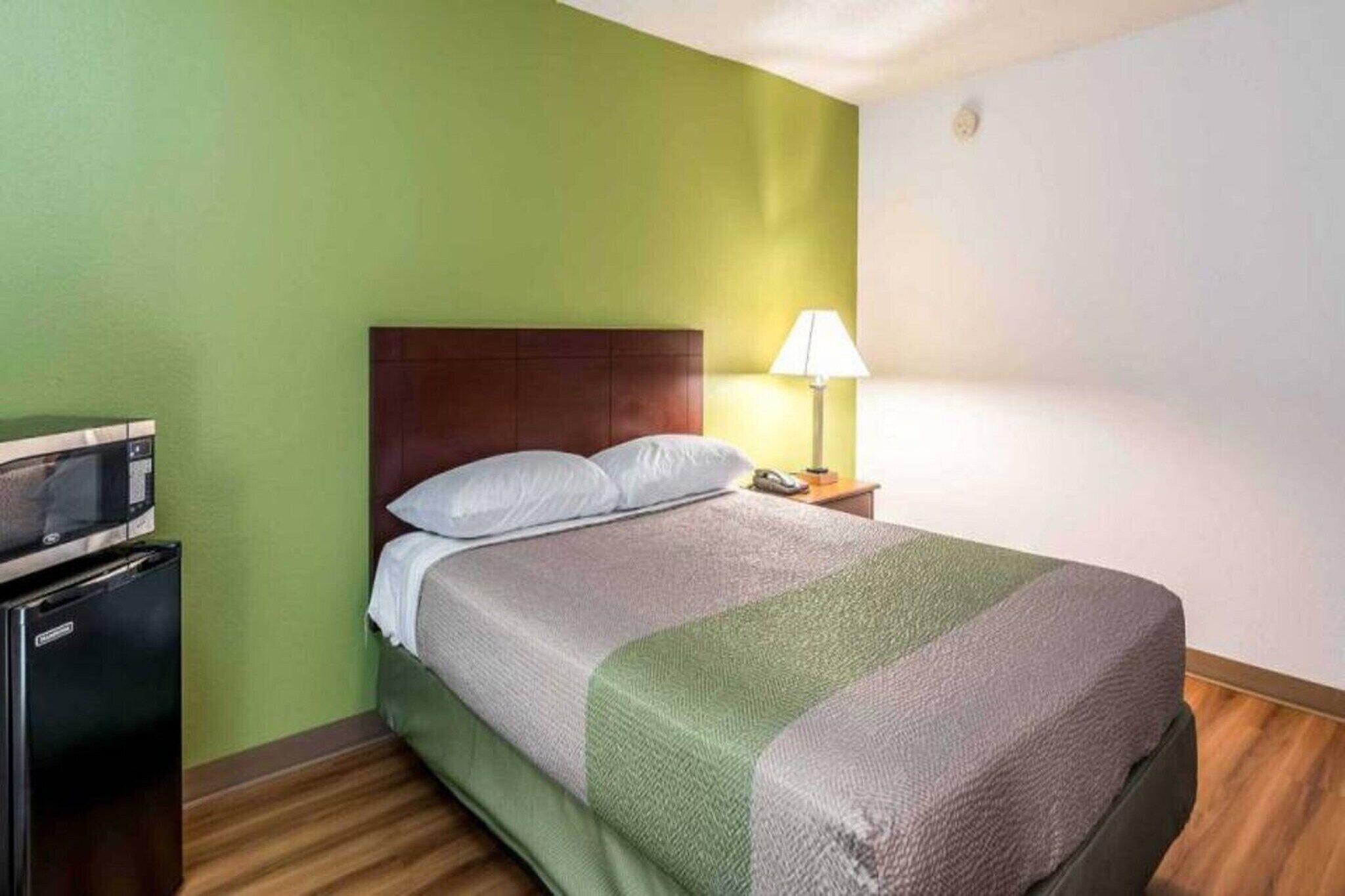 A hotel room with a bed, a nightstand with a lamp, and a kitchenette area.