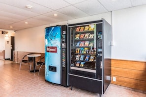 Máquina de vendas com lanches e dispensador de água numa área pública com chão em mosaico e um banco.