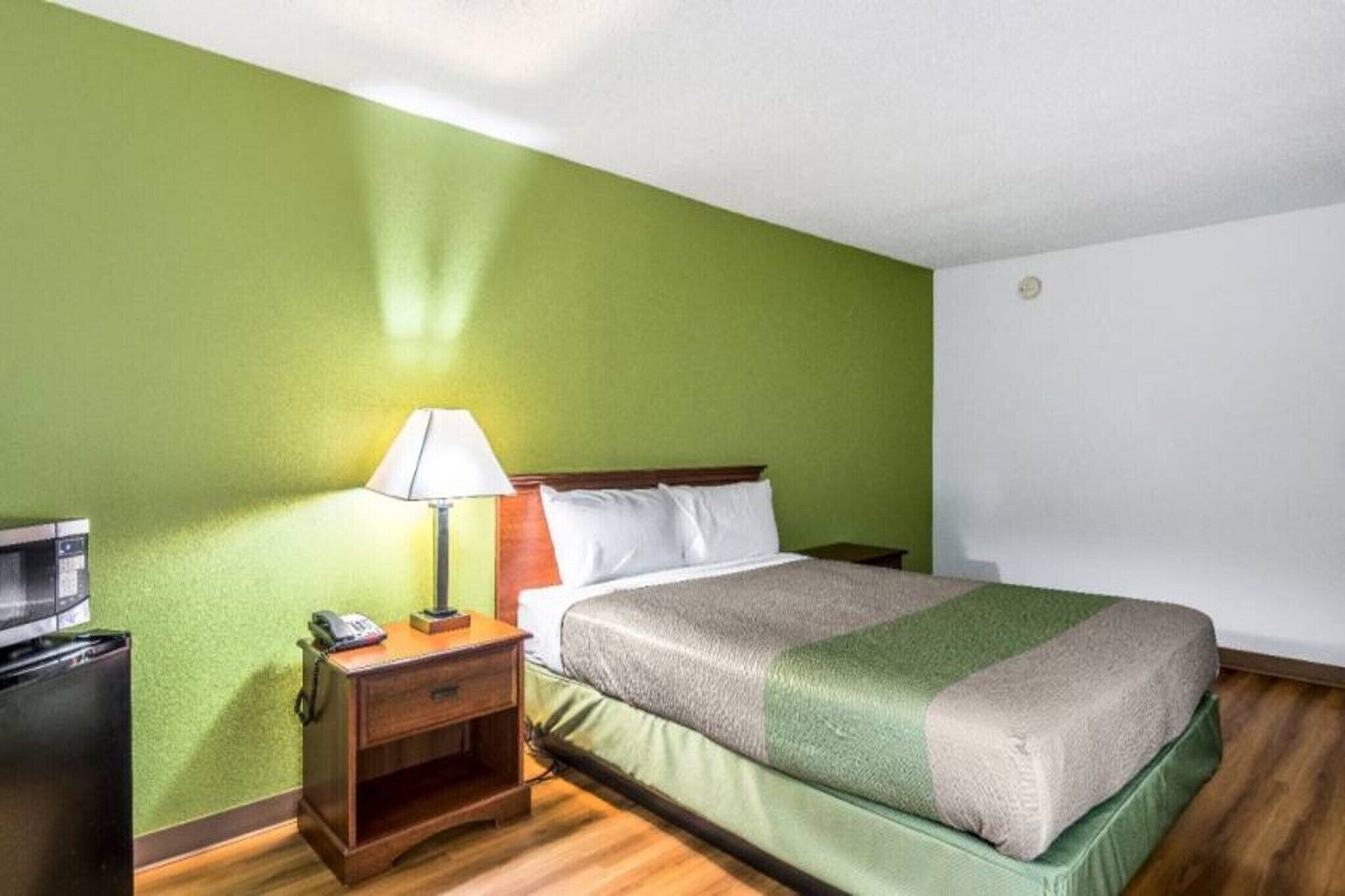 A hotel room with a bed, a nightstand with a lamp, a telephone, and a small desk with a computer.