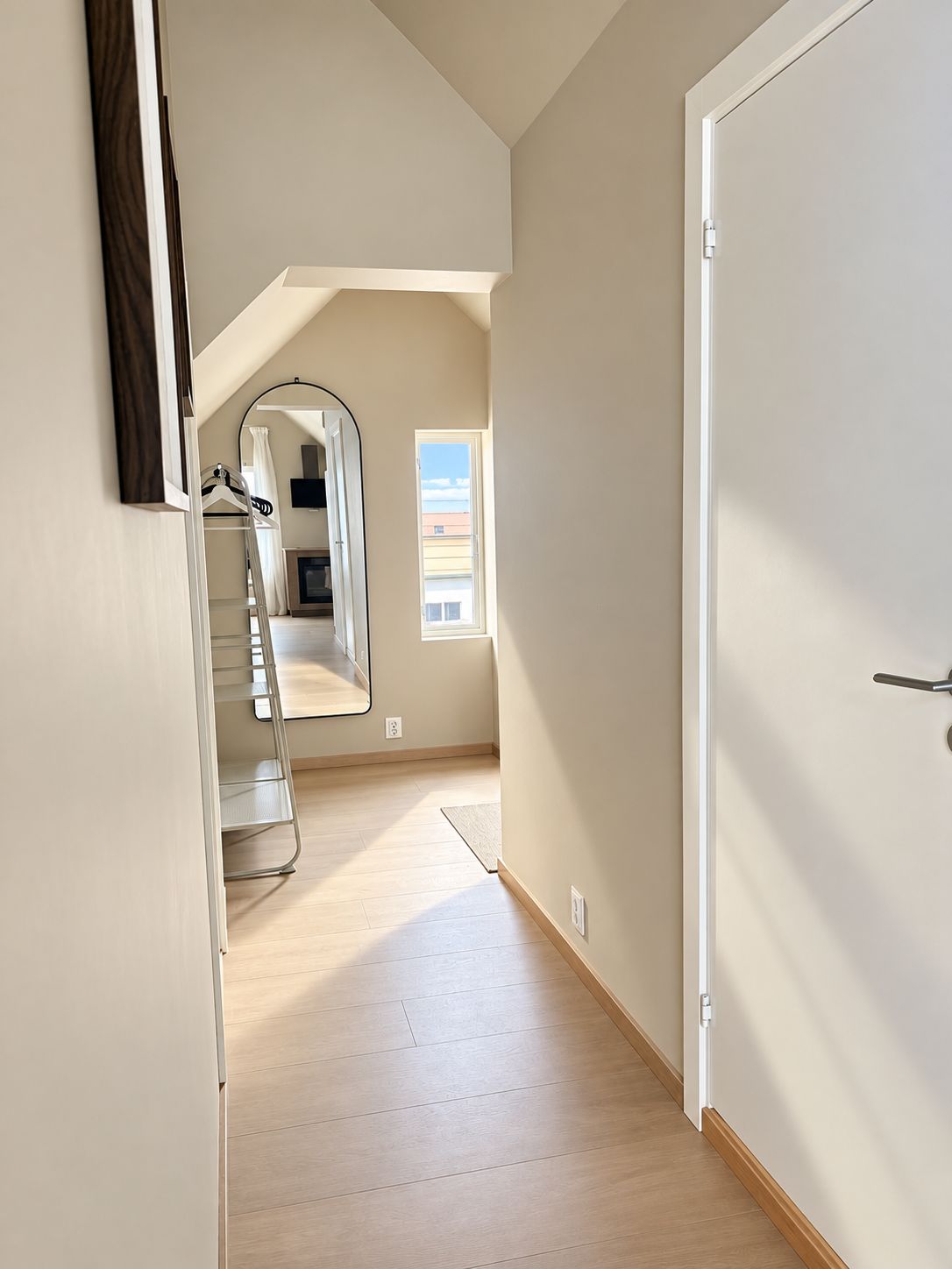 A hallway with a wooden floor, a white door, and a window with a view of buildings.