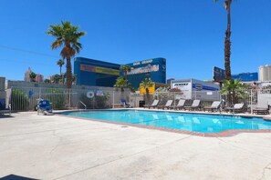Una piscina con lettini e un'area recintata, circondata da palme e edifici sullo sfondo.