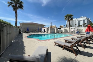 A swimming pool with lounge chairs and umbrellas, surrounded by a fence and palm trees.