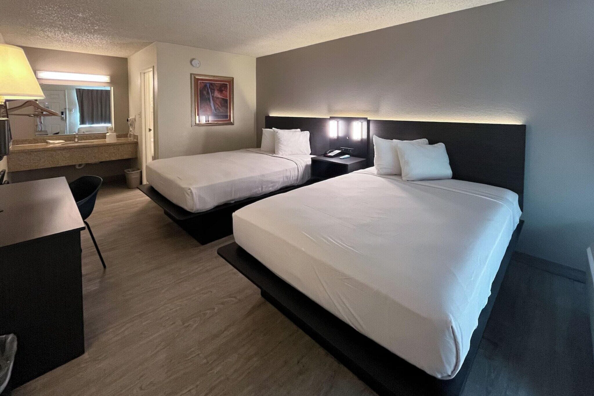 Une chambre d’hôtel avec deux lits, un bureau, une chaise, une table de chevet et une lampe fixée au mur.