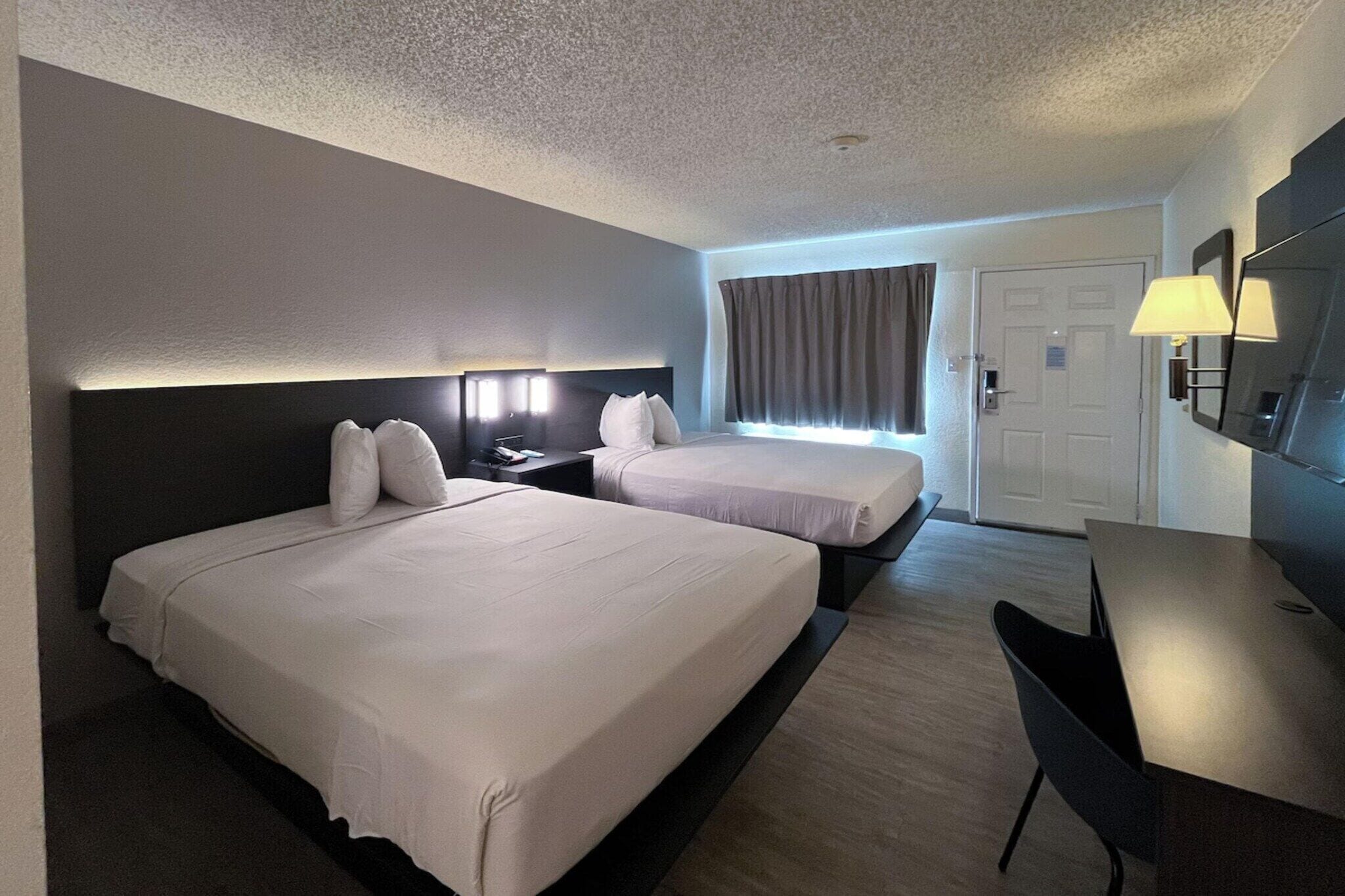 A hotel room with two beds, a desk, a chair, a TV, and a door.