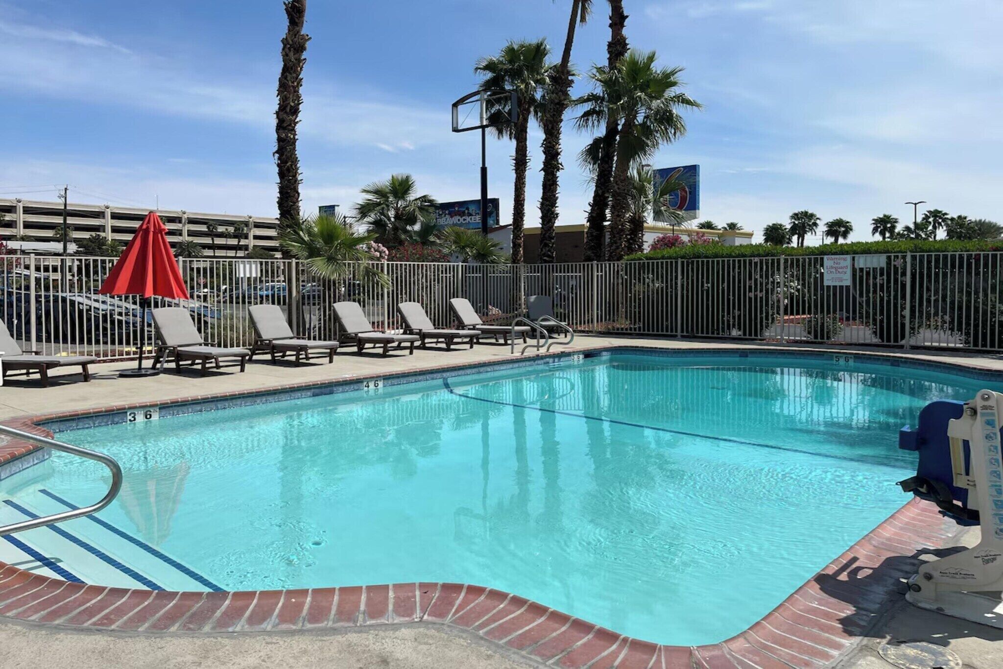 A swimming pool with lounge chairs and umbrellas, surrounded by palm trees and a fence.