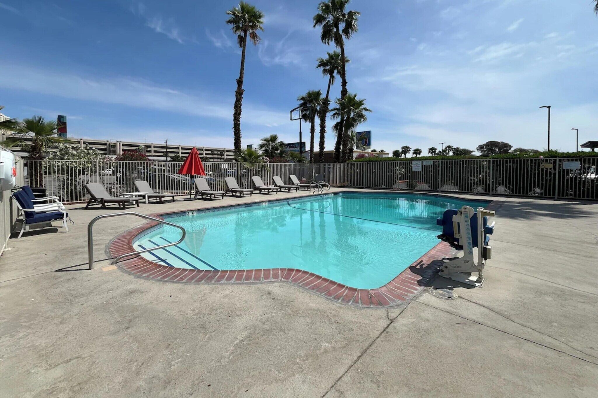 A swimming pool with lounge chairs and a red umbrella, surrounded by palm trees and a fence.