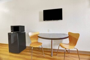Een kamer met een flatscreen televisie aan de muur, een kleine zwarte koelkast en twee houten stoelen rond een ronde tafel.