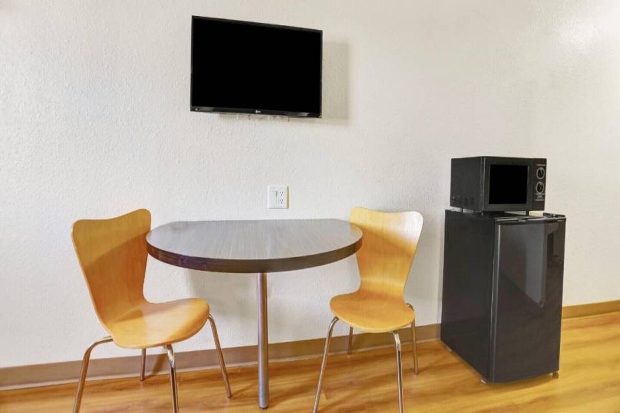 A room with a round table, two chairs, a television mounted on the wall, and a small black refrigerator.