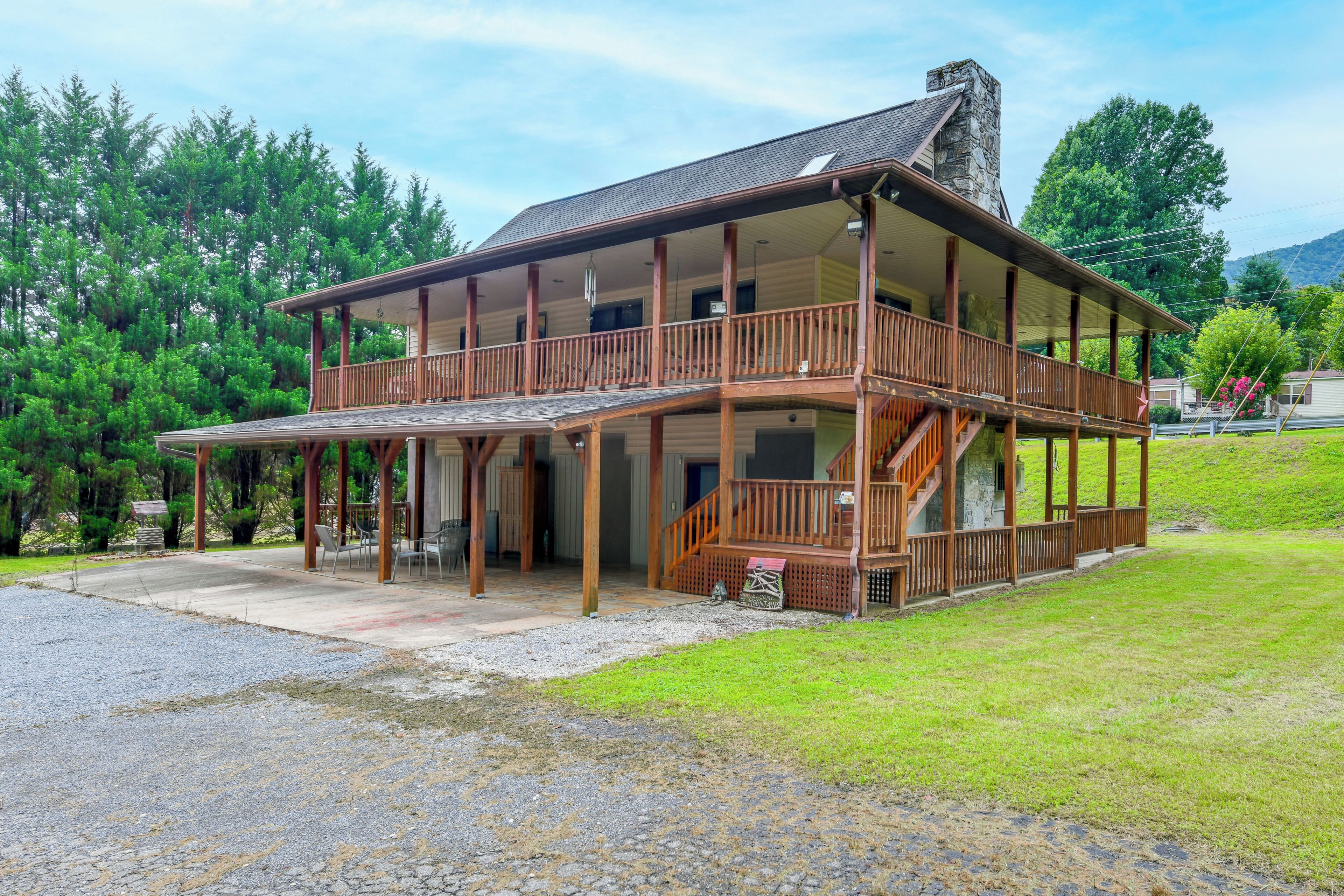A two-story wooden cabin with a balcony, surrounded by a gravel driveway and lush greenery.