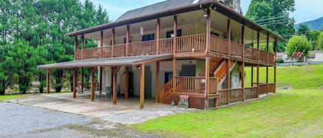 A two-story wooden cabin with a balcony, surrounded by a gravel driveway and lush greenery.