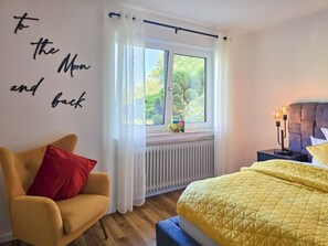 Ein Schlafzimmer mit Bett, Sessel, einem Fenster mit Vorhängen, Heizkörper und Nachttisch mit Lampe.