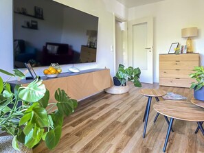 Ein Wohnzimmer mit Holzboden, einem Fernseher, einem Holzkasten, einem Esstisch und Topfpflanzen.