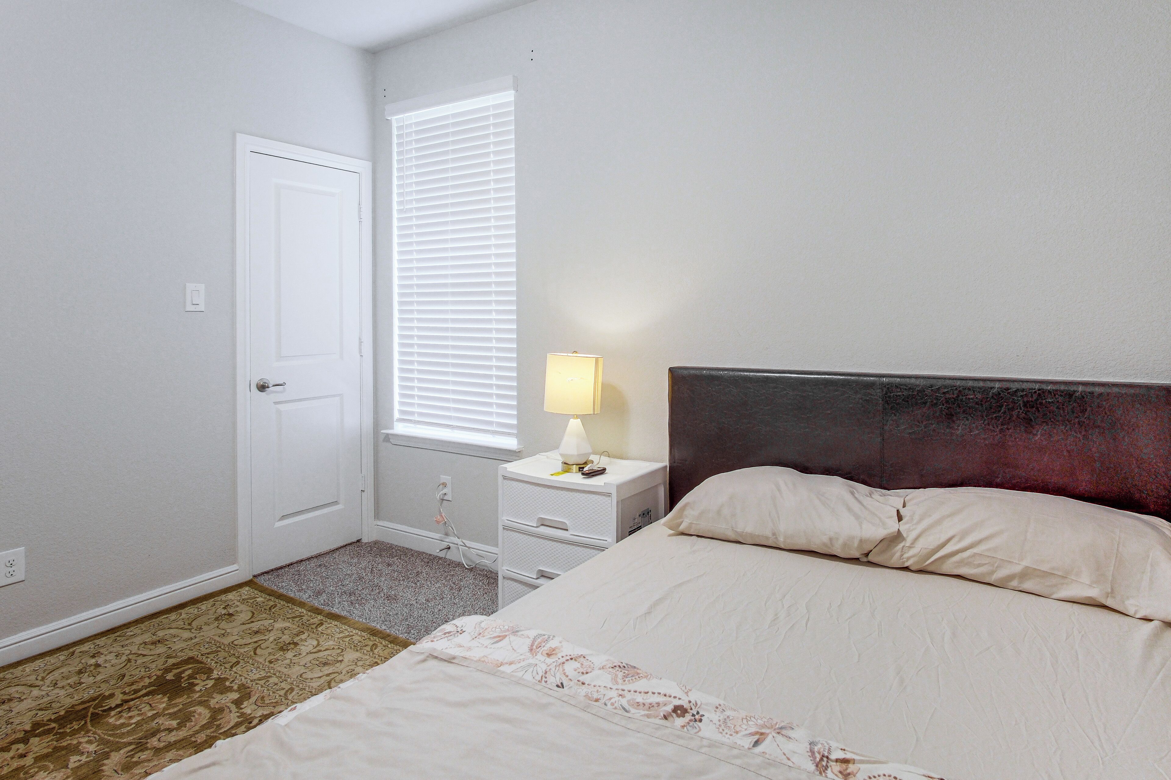 Une chambre d’hôtel avec un lit, une table de chevet, une lampe, une porte et une fenêtre avec des stores.