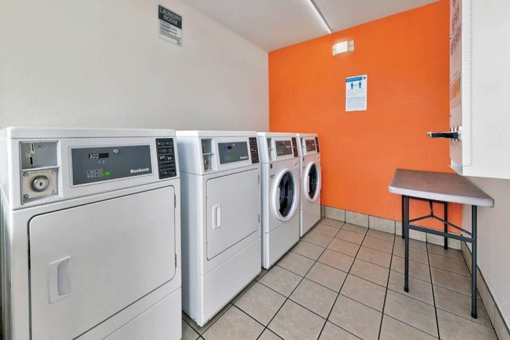 A laundry room with four washing machines, a small table, and an orange wall.