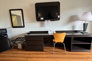 A compact hotel room with a desk, a chair, a television mounted on the wall, a lamp, and a small trash bin.