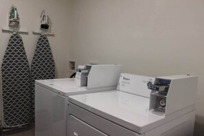 A laundry room with two white washing machines and a dryer, a wall-mounted ironing board, and a shelf with ironing supplies.