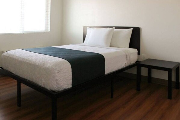 A single bed with a black frame, white bedding, and a dark-colored throw blanket. There is a small bedside table with a lamp. The room has wooden flooring and a window with blinds.