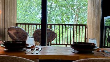 A dining area with a table set for two, a view of trees through large windows, and a wooden ceiling.