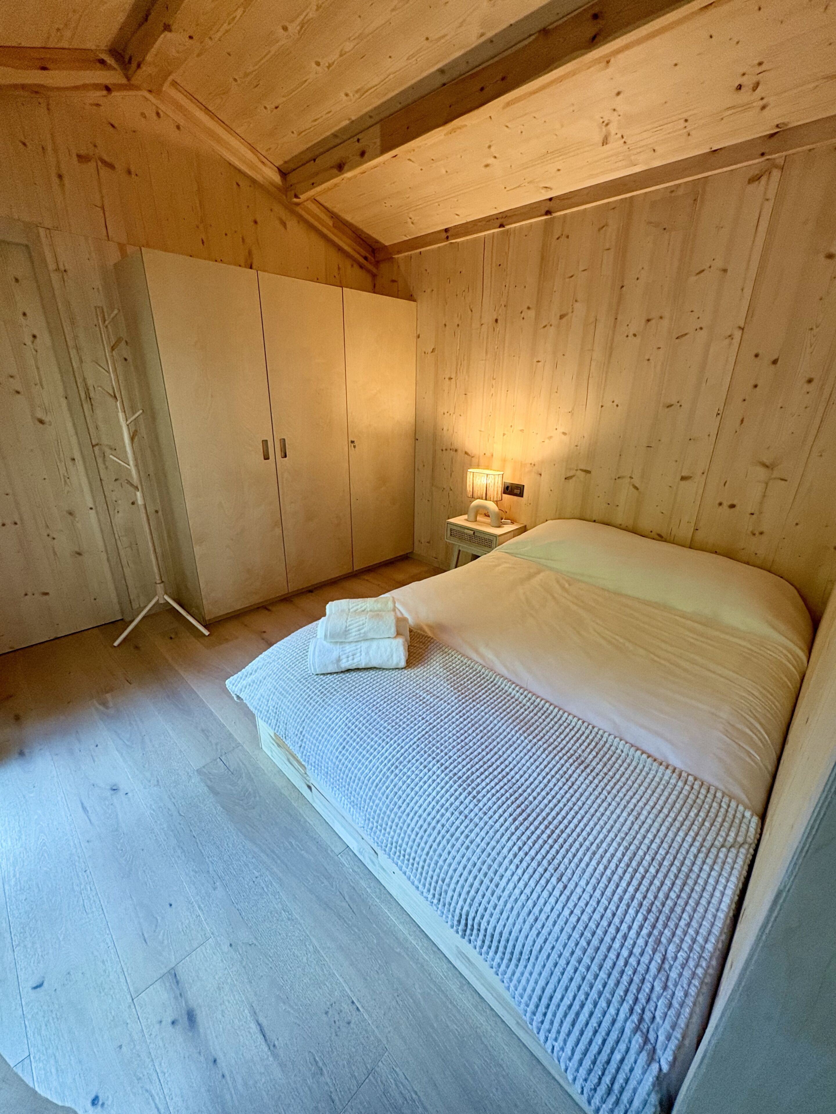 A wooden room with a bed, a wardrobe, and a nightstand with a lamp.