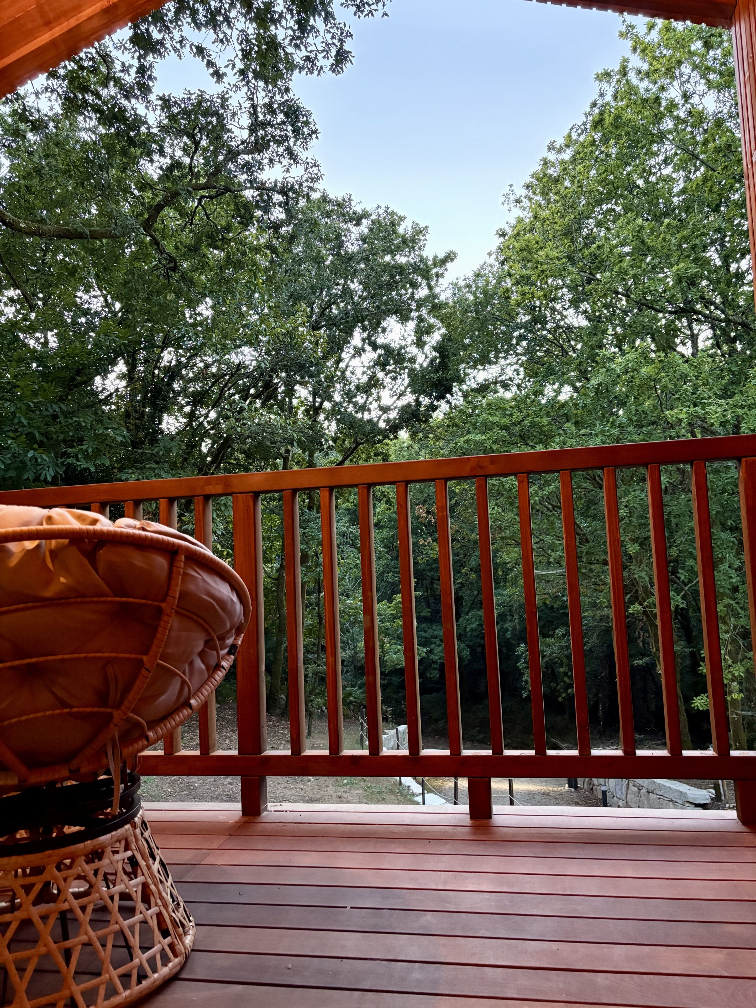A wooden deck with a railing, a wicker chair, and a view of dense green trees.