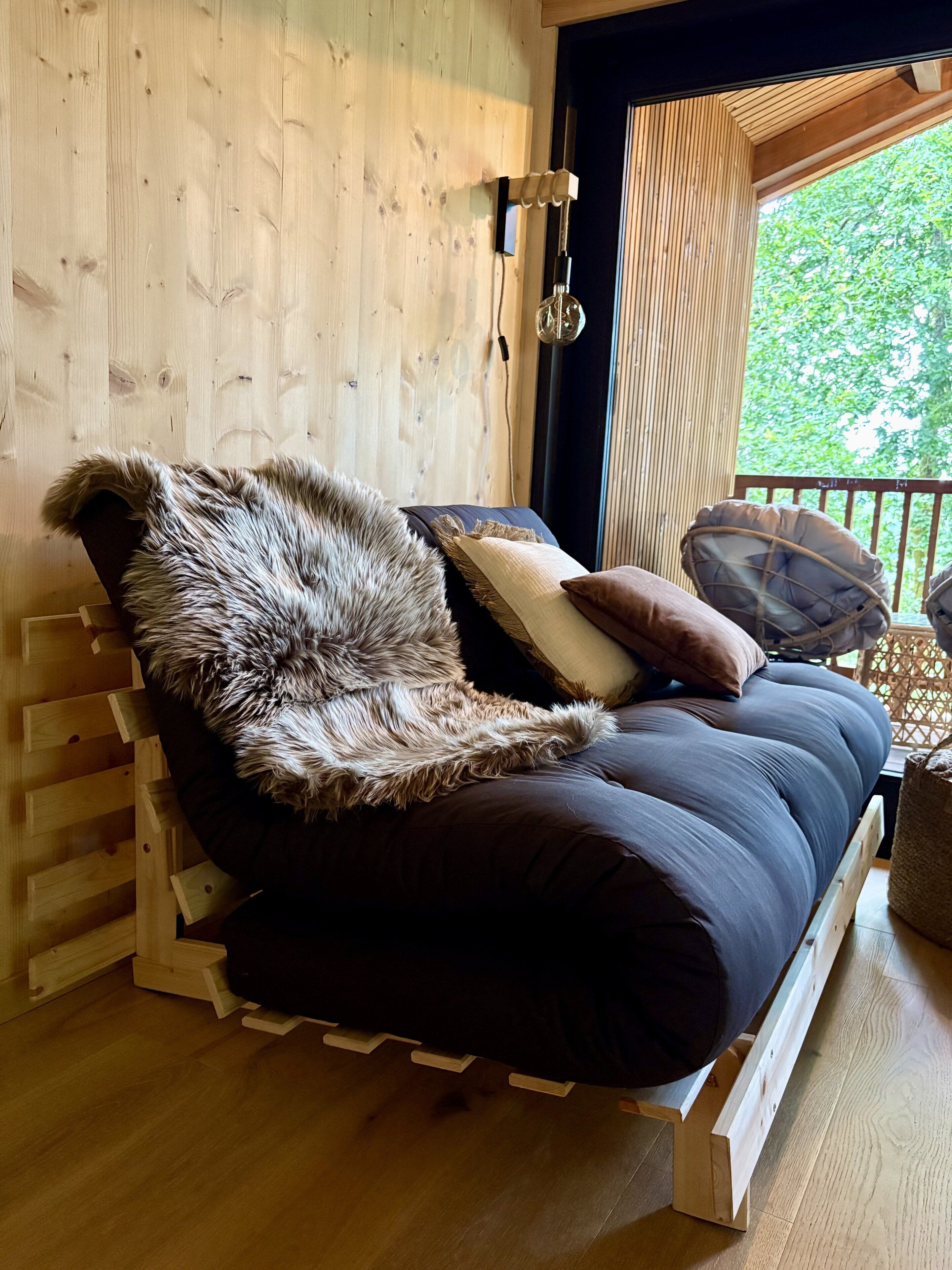 Interieur van een houten hutje met een bed, kussens en uitzicht op groen.