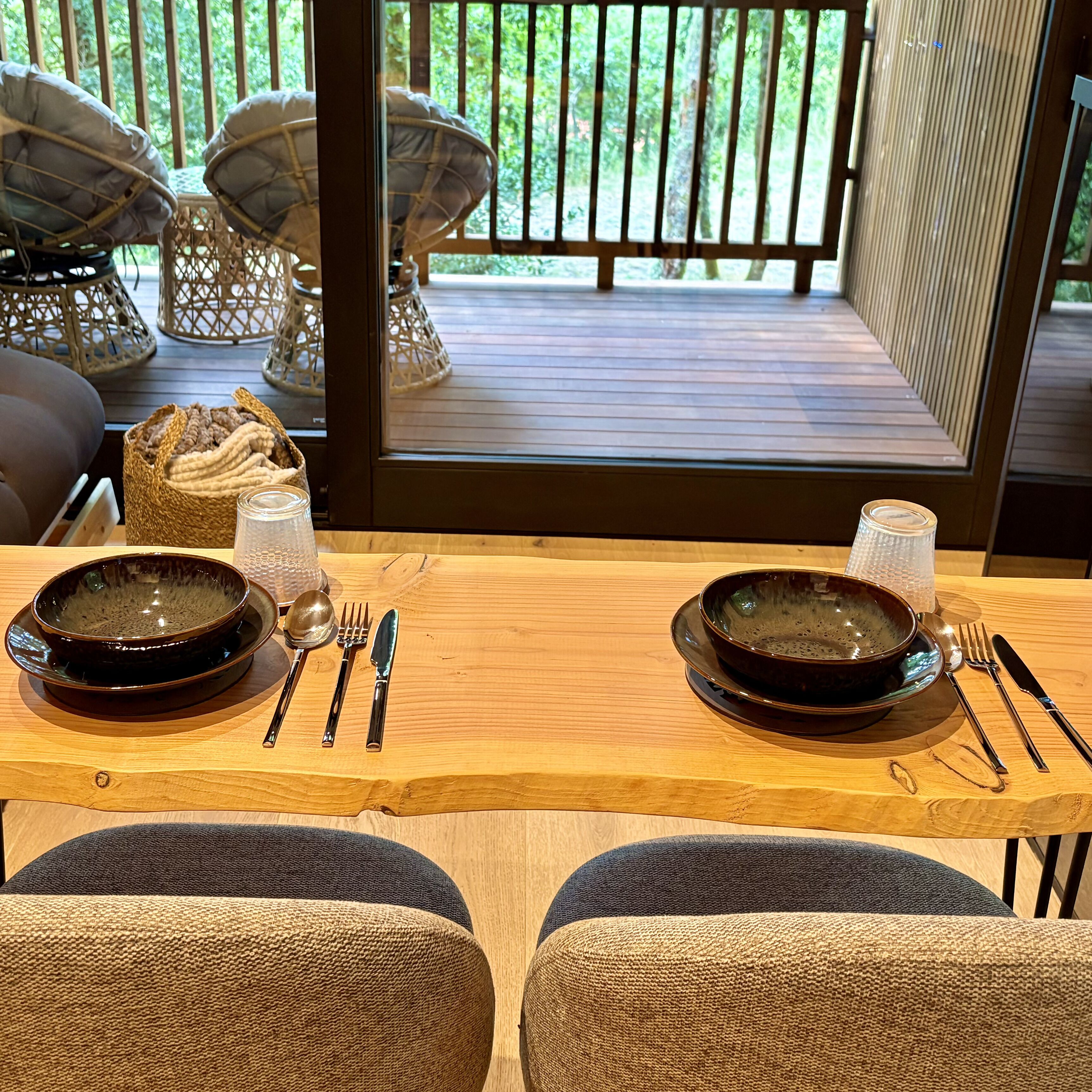 Mesa lista con platos, cubiertos y una cesta de pan, con vista a una terraza de madera con mobiliario exterior.