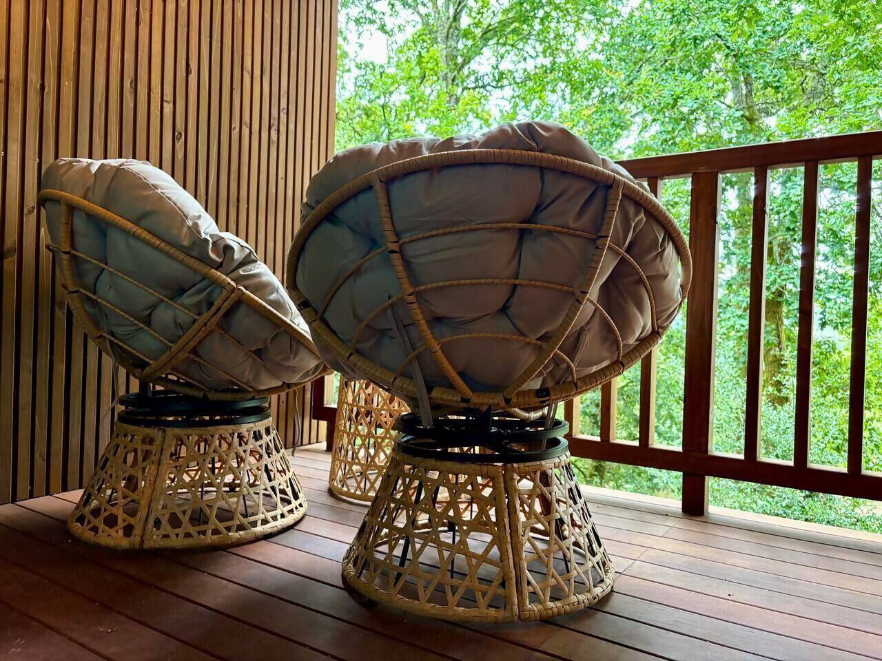 Twee rieten stoelen met kussens op een houten terras, met uitzicht op groen.