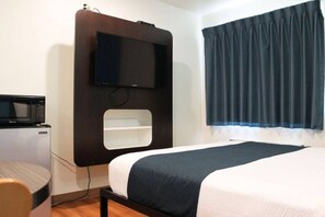 A hotel room with a bed, a television mounted on the wall, a small desk with a chair, and a window with curtains.