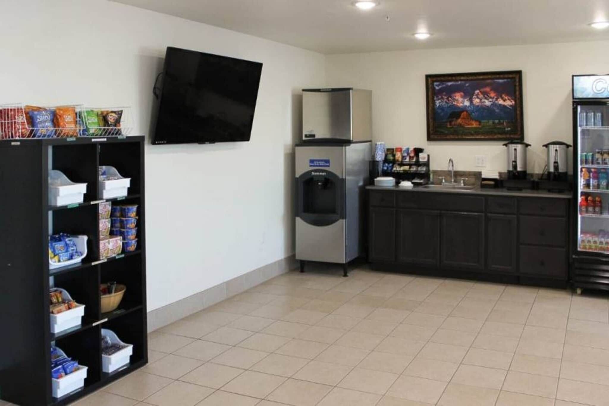 A clean, well-lit room with a flat-screen TV mounted on the wall, a refrigerator, a water dispenser, and a coffee machine on the counter.