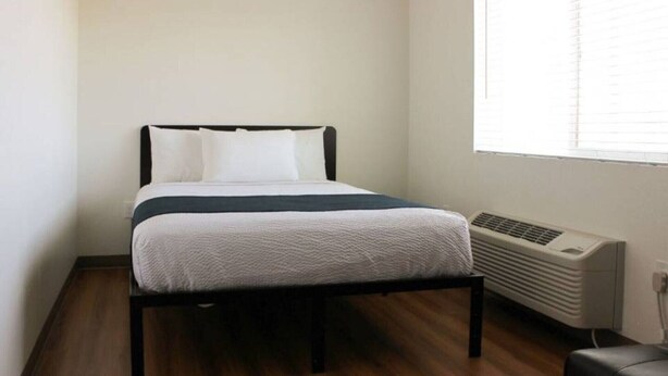 A single bed with a dark frame, white bedding, and a window with blinds to the right.